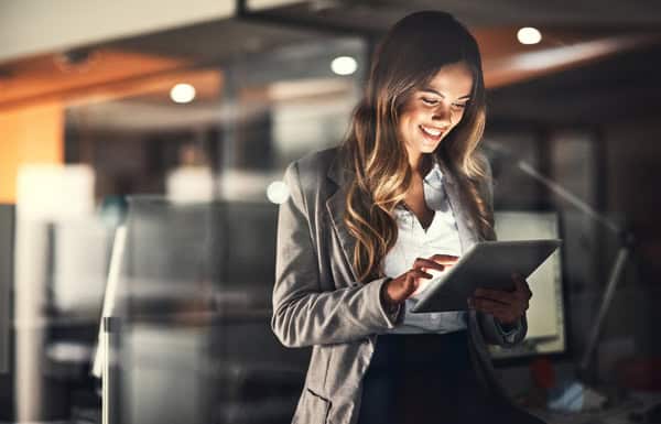 Eine Frau in einem Geschäftsanzug steht in einem Büro, konzentriert auf ein Tablet in ihren Händen, mit einem modernen Arbeitsbereich im Hintergrund.