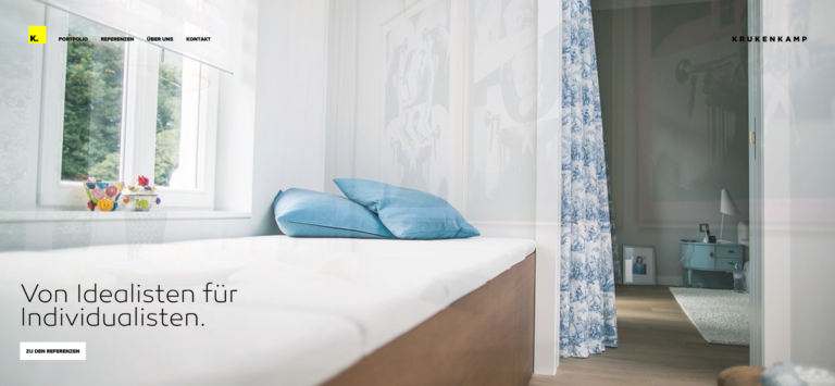 Helles, minimalistisches Schlafzimmer mit einem Bett mit blauen Kissen, geblümten Vorhängen und natürlichem Licht durch ein Fenster.